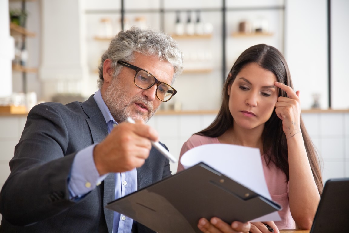 Serious mature legal advisor reading, analyzing and explaining document to female customer. Man and woman sitting at table and reading papers. Expertise and paperwork concept